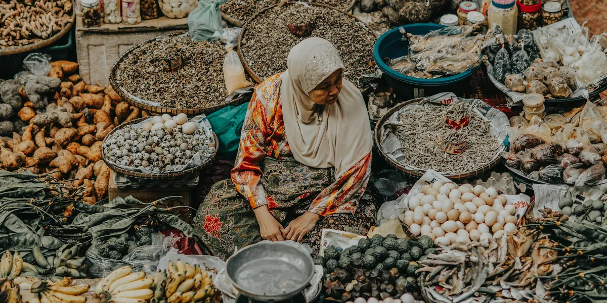 A trader in a headscarf surrounded by baskets of dried goods, eggs and produce at a market.