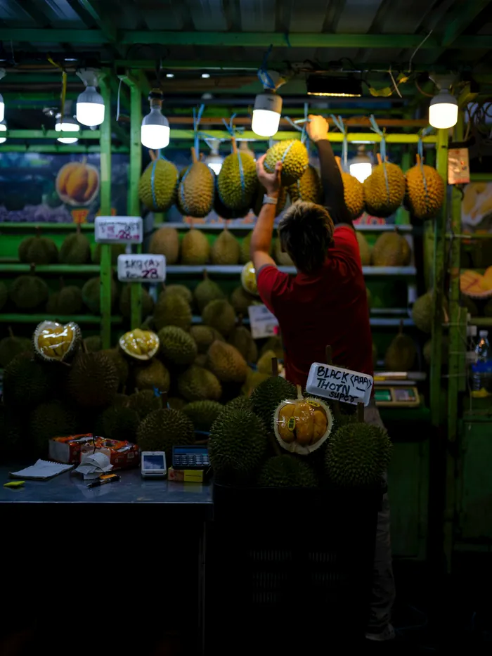 A vendor arranging durians on racks under strip lights at a night market stall.