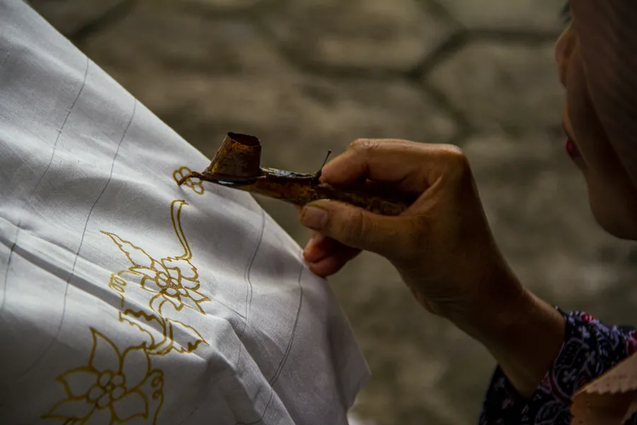 A hand drawing wax patterns onto white fabric with a canting tool.