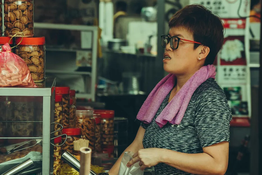 A shopkeeper in glasses working behind a counter stacked with jars of dried goods.