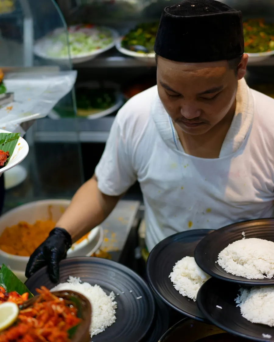 A cook in a songkok plating rice and curry onto black plates at a food stall.