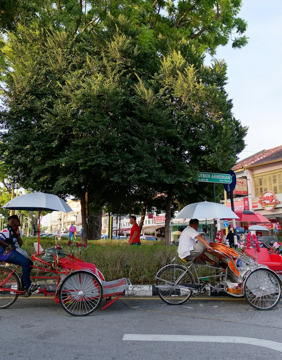 Trishaw riders waiting under a rain tree on a George Town street in Penang.