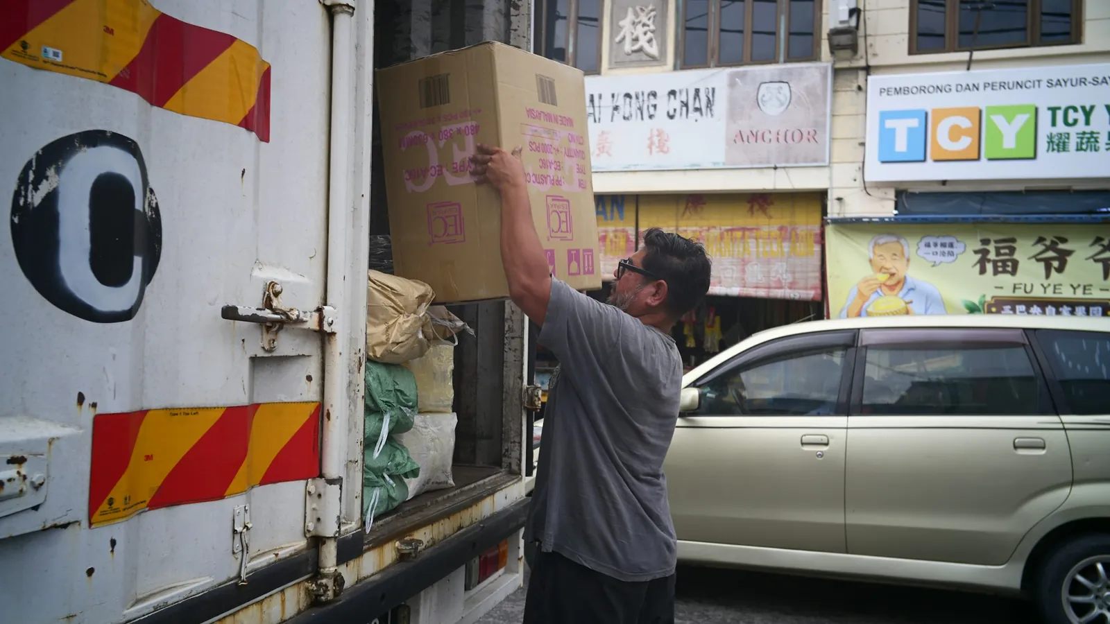 A man loading a cardboard box into a delivery van on a shophouse street.
