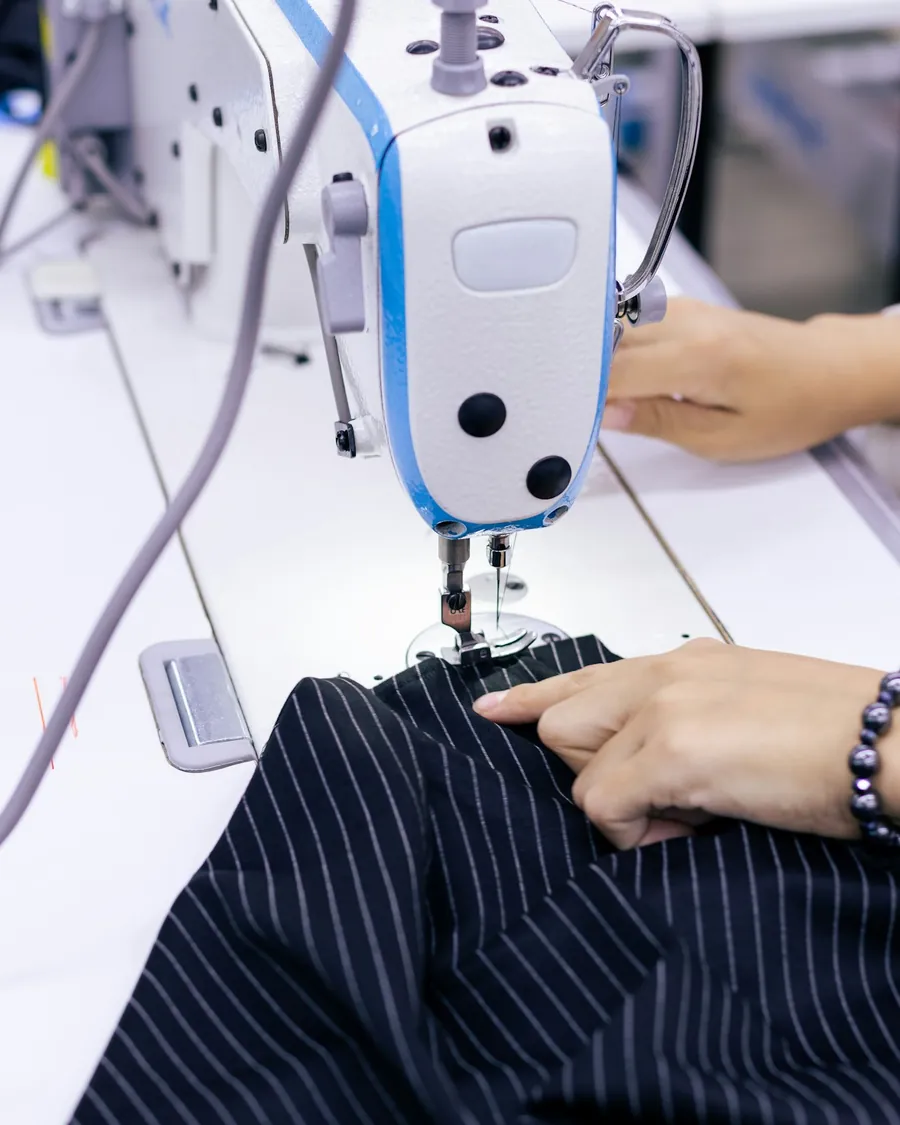 Hands guiding pinstriped fabric through an industrial sewing machine.